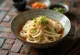 Kišimen udon, postrežen v skledi s ploskimi udon rezanci v bogati daši osnovi, okrašen z mlado čebulo in sezamovimi semeni